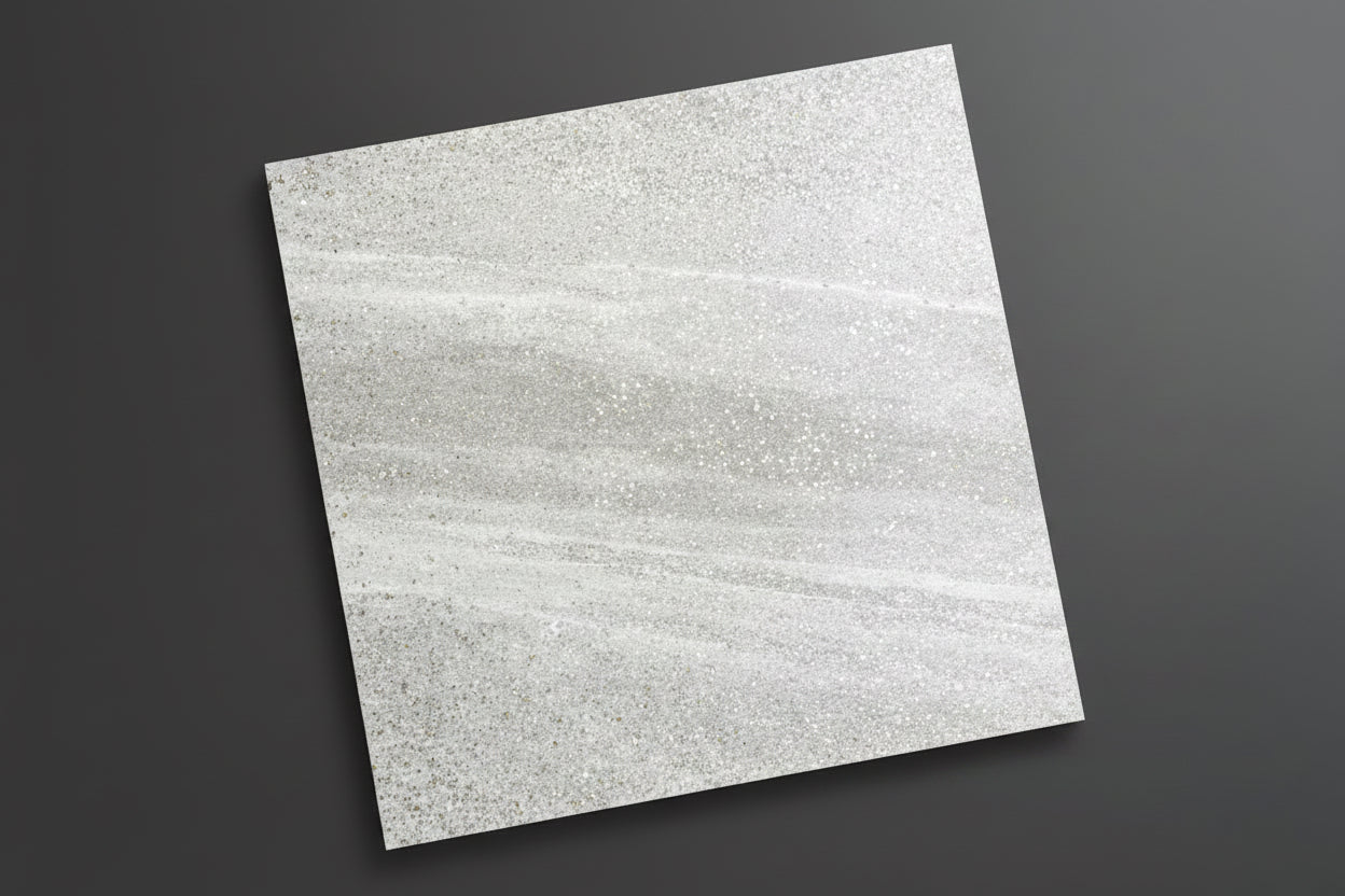 A square gray stone-look tile with a subtle white veining pattern lies on a beige tiled floor. A person's legs and feet wearing black and white striped slide sandals are visible at the bottom right, indicating a lifestyle or in-situ product presentation.