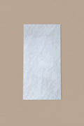 A large rectangular white marble-look tile or panel with subtle gray veining, shown standing on a beige tiled floor next to a person's legs in blue jeans and brown sandals.