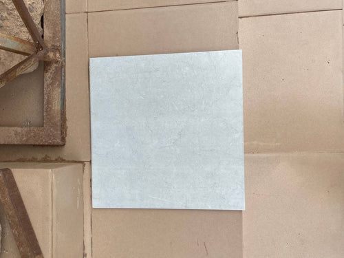 A square, light gray textured tile or stone sample lies on a tan tiled floor, with a person's feet in brown sandals visible on the right edge.