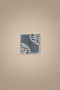 A square decorative tile with a dark gray center and mosaic stone accents in gray, brown, and light blue, placed on a beige tiled floor. A person's feet wearing brown sandals and blue jeans are visible at the bottom of the frame.
