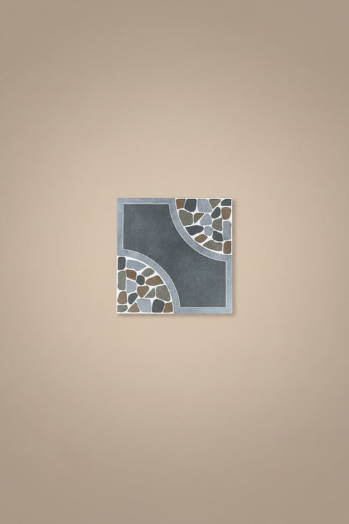 A square decorative tile with a dark gray center and mosaic stone accents in gray, brown, and light blue, placed on a beige tiled floor. A person's feet wearing brown sandals and blue jeans are visible at the bottom of the frame.